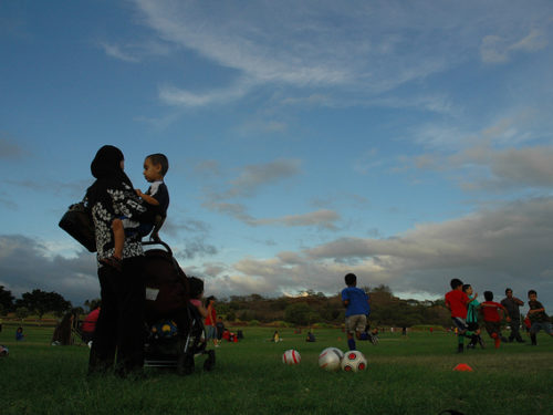 Mom holding kid watching kids playing a soccer game